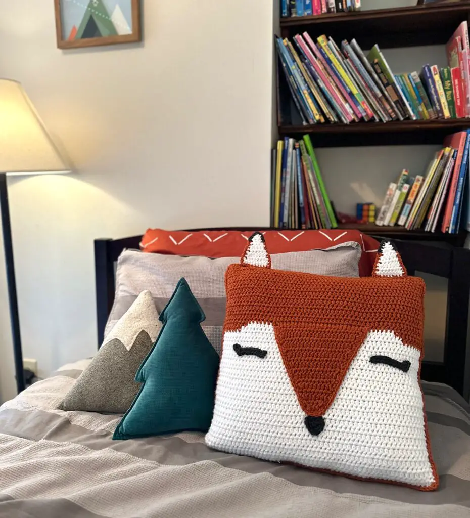 an orange crocheted fox pillow sits on a gray bed in a nursery.