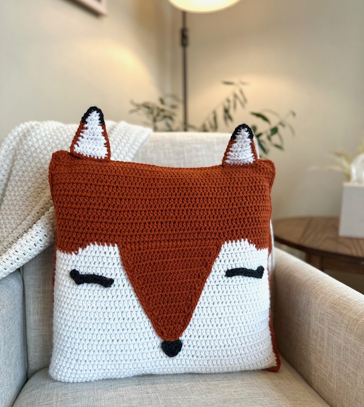 a crochet orange and white fox pillow sites on a white chair.