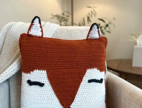 a crochet orange and white fox pillow sites on a white chair.